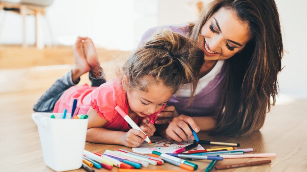 mãe e filha sorrindo e deitadas no chão segurando canetinhas hidrocor para fazer uma atividade lúdica