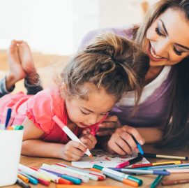 mãe e filha sorrindo e deitadas no chão segurando canetinhas hidrocor para fazer uma atividade lúdica